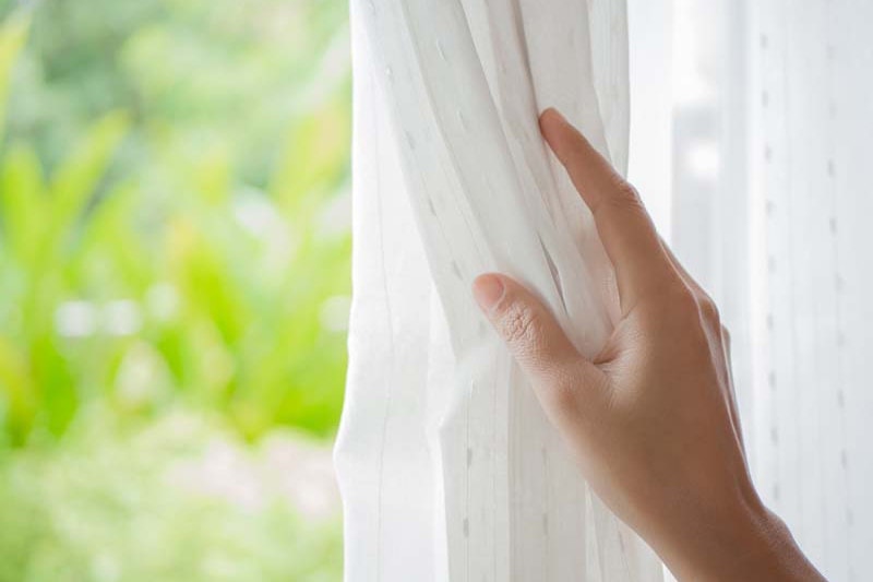 A hand pulling open a while curtain to reveal greenery outside.